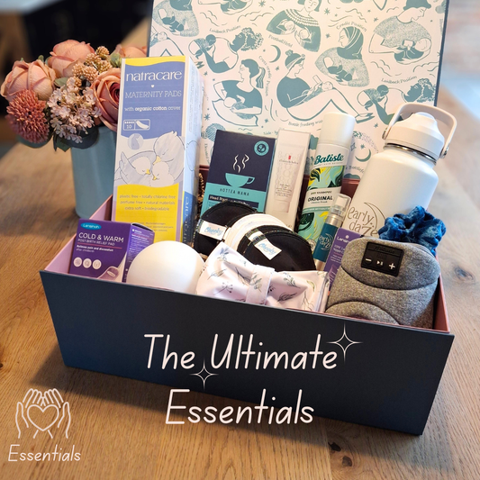 Box labeled 'The Ultimate Essentials' with various postpartum gift items including a water bottle, tea and other products on a wooden surface.